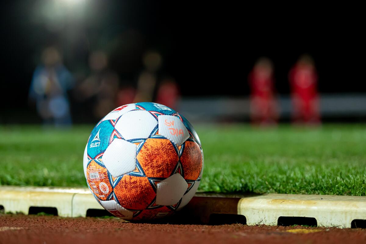 Trainingsball in Wei&szlig;, Blau und Orange. Im Hintergrund trainieren Spieler bei Flutlicht